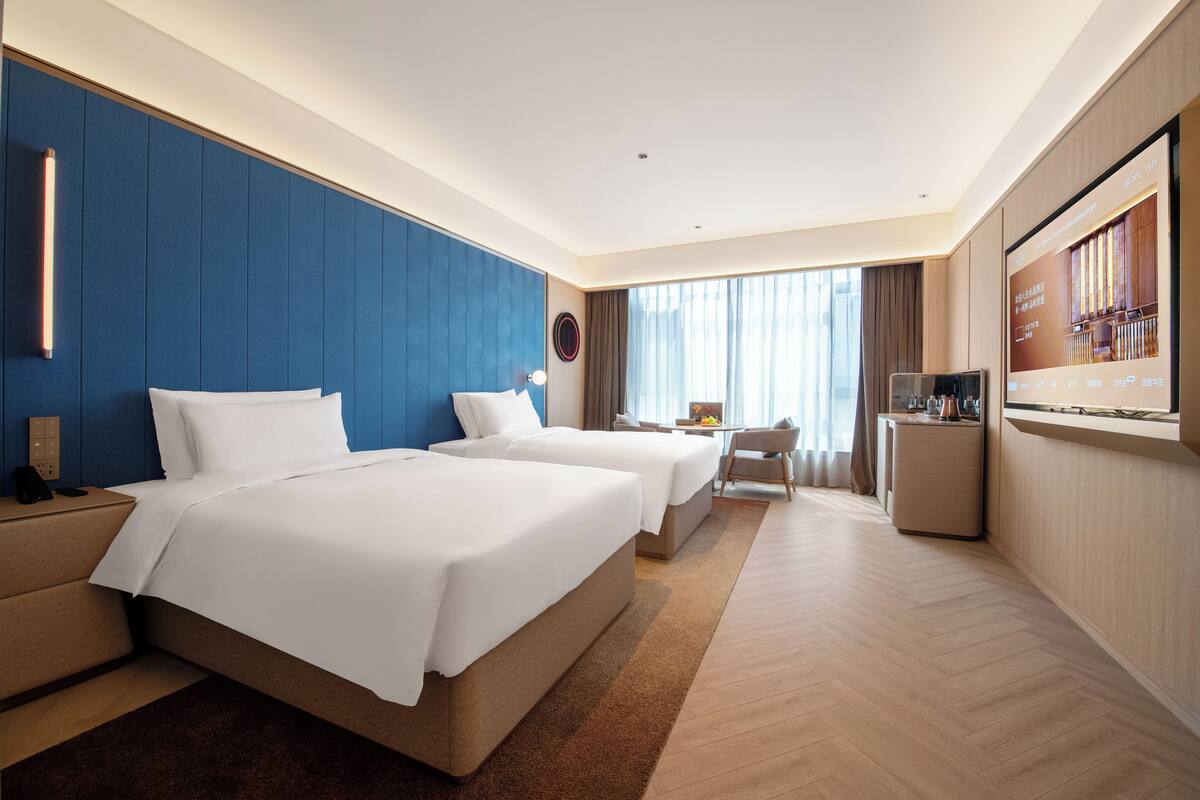 A hotel room with two beds, a desk, a TV, and a large window.