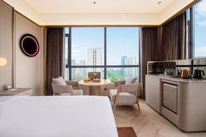 A hotel room with a bed, a dining table with chairs, a kitchen area, and large windows offering a city view.