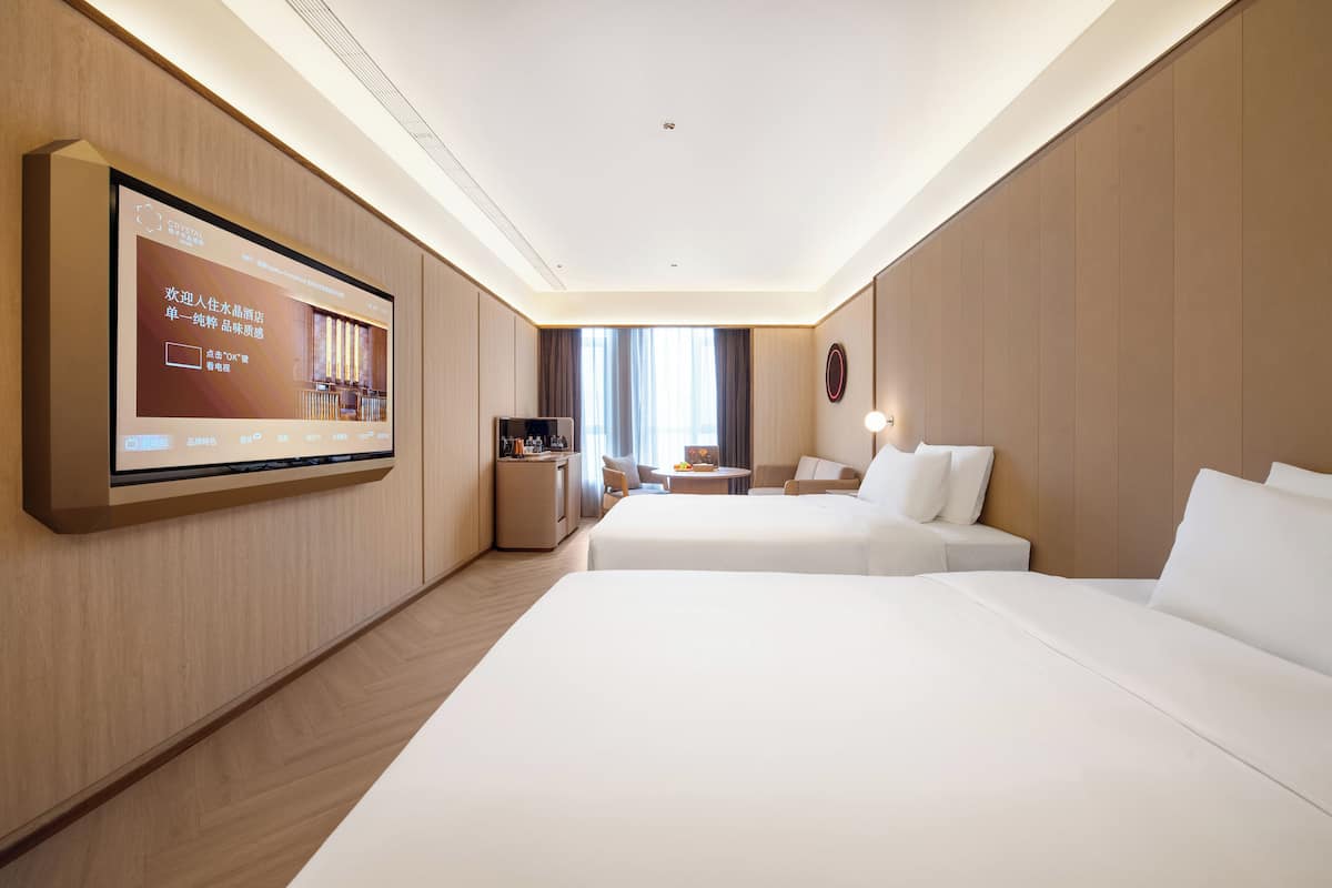 A hotel room with a large flat-screen TV mounted on the wall, two beds, a small desk with a chair, and a window with curtains.