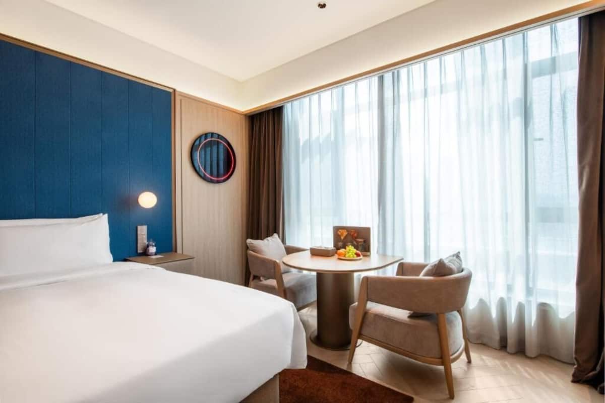 A hotel room with a large bed, a round table with chairs, a wall clock, and a large window with curtains.