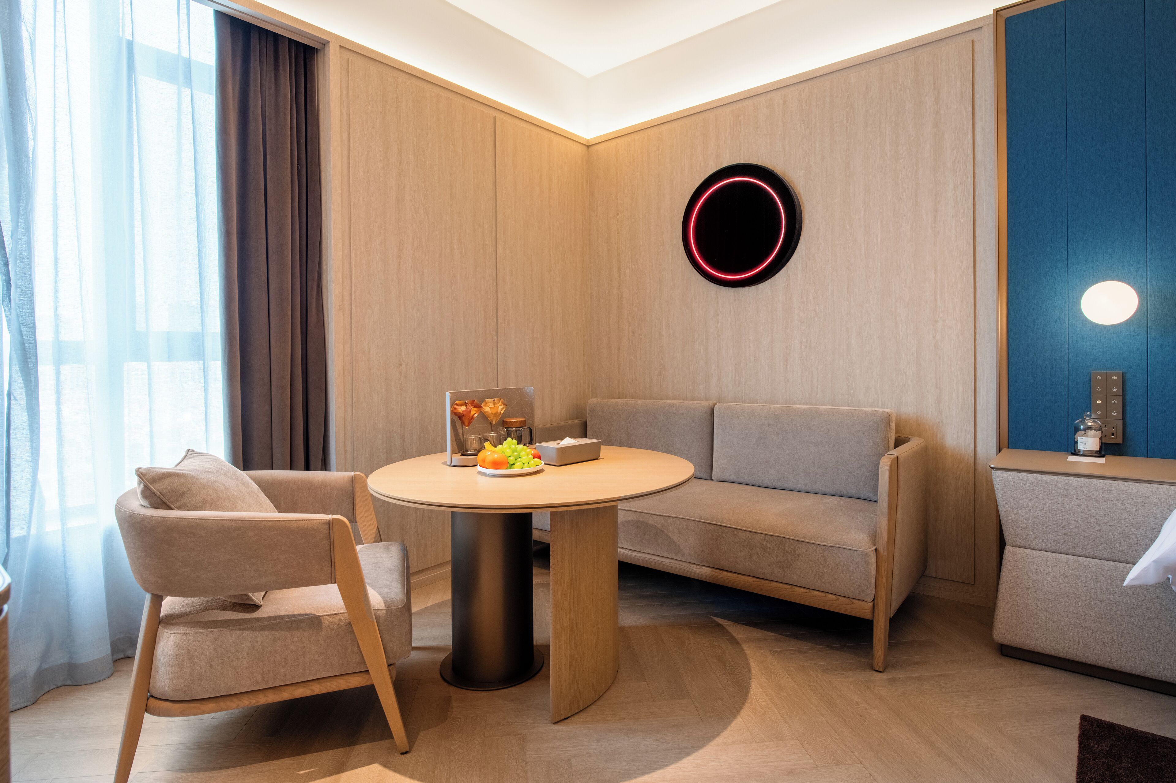 A modern hotel room with a round table, two sofas, a coffee table, a wall-mounted circular light, and a view of the outdoors.