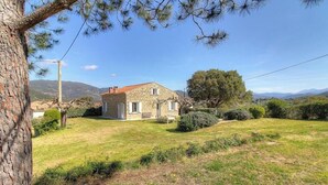 Une maison en pierre avec un toit rouge, entourée d’une cour verdoyante et d’arbres.