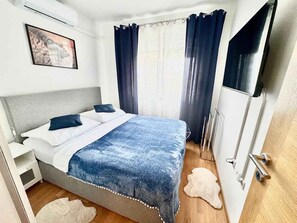 Ein Schlafzimmer mit einem Bett, einem weißen Nachttisch, einem flauschigen weißen Teppich, Holzboden, einem Fenster mit Vorhängen und einer Wand-Klimaanlage.