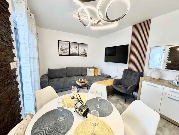 Ein modernes Wohnzimmer mit einer Couch, einem Esstisch mit Platzsets, einem an der Wand montierten Fernseher und einer Holzkassette.