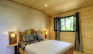 Un dormitori de cabana de fusta amb un llit, dues làmpades penjades a la paret, una finestra amb cortines i vista a arbres.