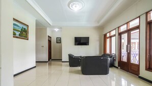 Un hall d’entrée spacieux, doté d’un sol carrelé, d’un téléviseur à écran plat fixé au mur et d’un coin salon avec des fauteuils noirs.