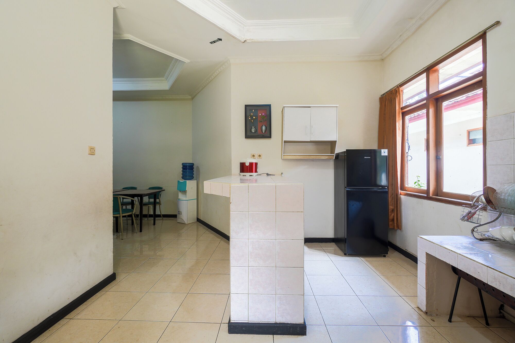 A compact kitchen with a refrigerator, a water dispenser, a dining table with chairs, and a window with curtains.