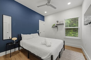 Ein Schlafzimmer mit einem Bett, einem Nachttisch, einem Deckenventilator, einem Fenster mit Jalousien und einem an der Wand befestigten Regal.