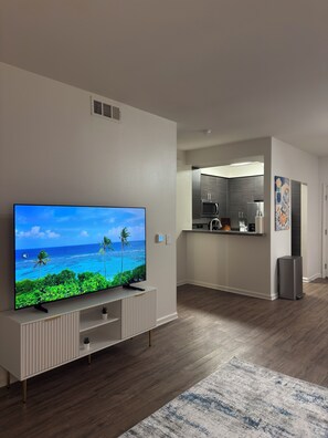 A modern living room with a flat-screen TV, a kitchen with dark wood flooring, and a hallway leading to a door. - Elegant 1-bedroom apartment in tranquil Marina del Rey (Marina del Rey)