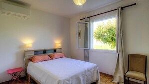 Ein Schlafzimmer mit einem Bett, Nachttischlampen, einem Sessel, einem Fenster mit Vorhängen und einer Klimaanlage.