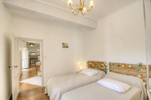 Ein Schlafzimmer mit zwei Betten, einem Holz-Kopfteil, einer Chandelüster und einem Flur, der zu einer Küche führt.