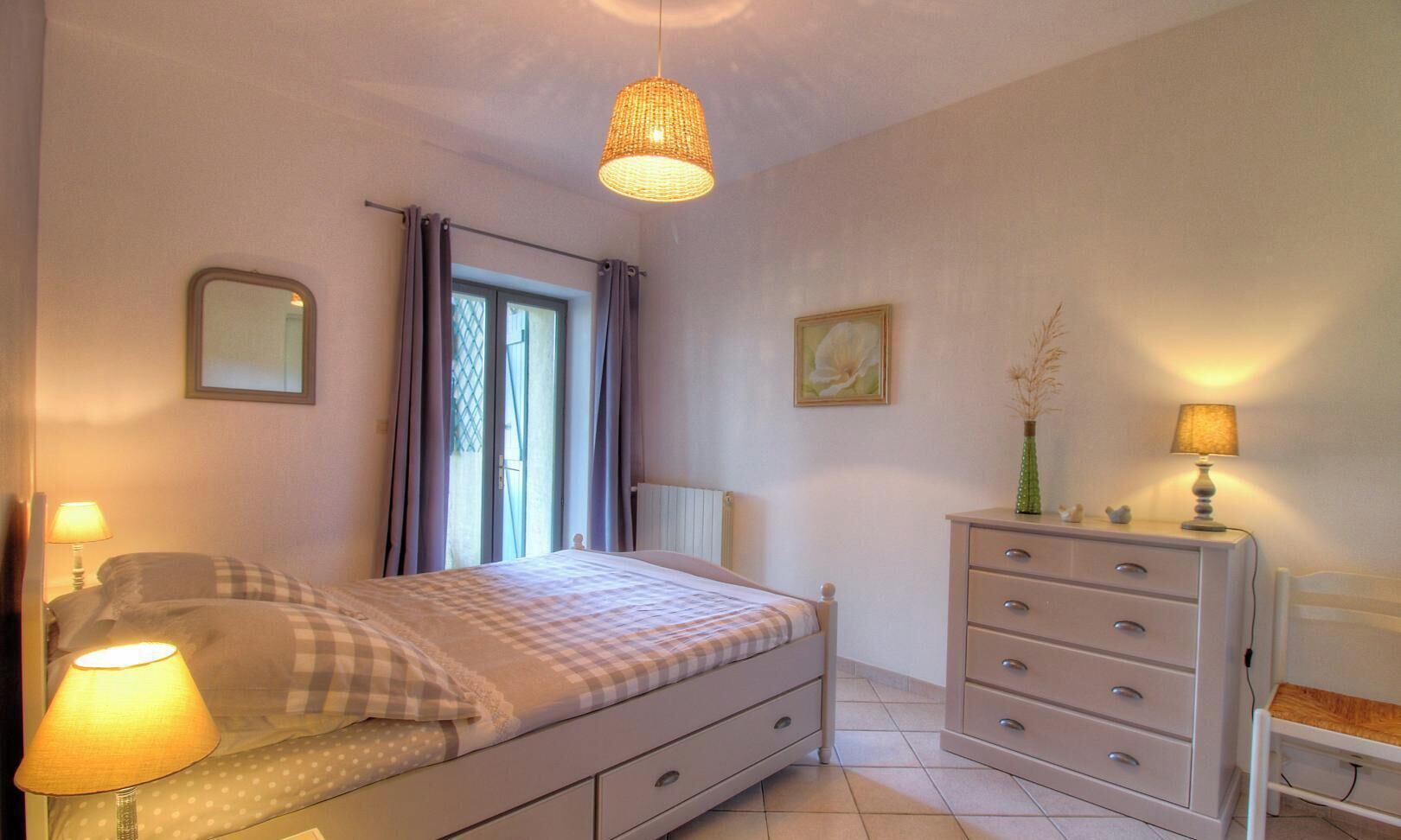 Un dormitorio con cama, lámparas de noche, cómoda, espejo y ventana con cortinas.