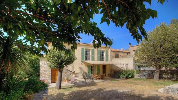 Una casa de dues plantes amb façana de pedra, persianes verdes i un porxo cobert amb mobles d'exterior.