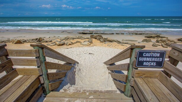 Ein Holzsteg führt zu einem Strand, mit einem Warnschild wegen versenkter Felsen.