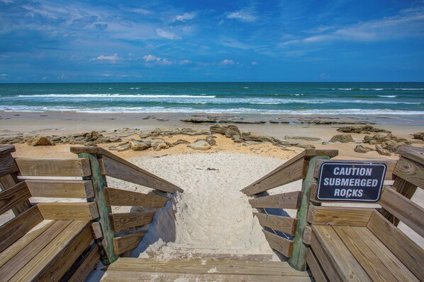 Ein Holzsteg führt zu einem Strand, mit einem Warnschild wegen versenkter Felsen.