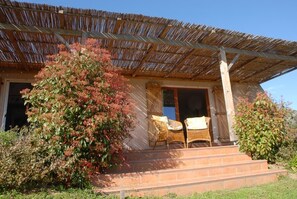 A building with a thatched roof, a porch with wicker furniture, and a large bush in front. - Rosemary - MONACIA D'AULLENE (MONACIA D'AULLENE)