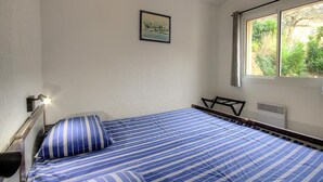 Ein Einzelbett mit einer blau-weißen gestreiften Tagesdecke, ein Nachttisch mit einer Lampe, ein Bild an der Wand und ein Fenster mit Blick auf Grünflächen.