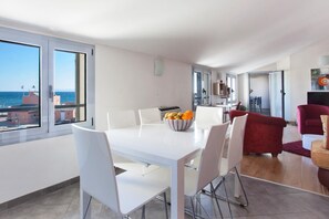 Un espace repas moderne avec une table et des chaises blanches, un saladier rempli de fruits et de grandes fenĂȘtres offrant une vue sur la mer et les bĂątiments.