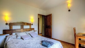 Une chambre avec une tête de lit en bois, un lit recouvert d’une literie à motifs, deux appliques murales, une porte en bois et une chaise.