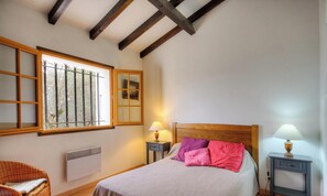 A bedroom with a bed, two bedside lamps, a window with a view, a radiator, and a wicker chair. - I caseddu d'Ulmetu 4 (OLMETO)