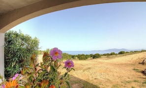 Blick durch ein rundes Fenster auf einen unbefestigten Weg, im Vordergrund violette Blumen und in der Ferne eine Berglandschaft.