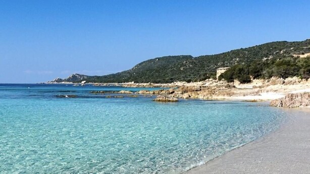 Ein Strand mit klarem Wasser, sandigem Ufer und einer hügeligen Landschaft im Hintergrund.