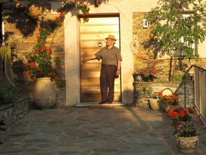 Un hombre parado frente a una entrada de piedra con una puerta de madera, rodeado de plantas en macetas y un muro de piedra.