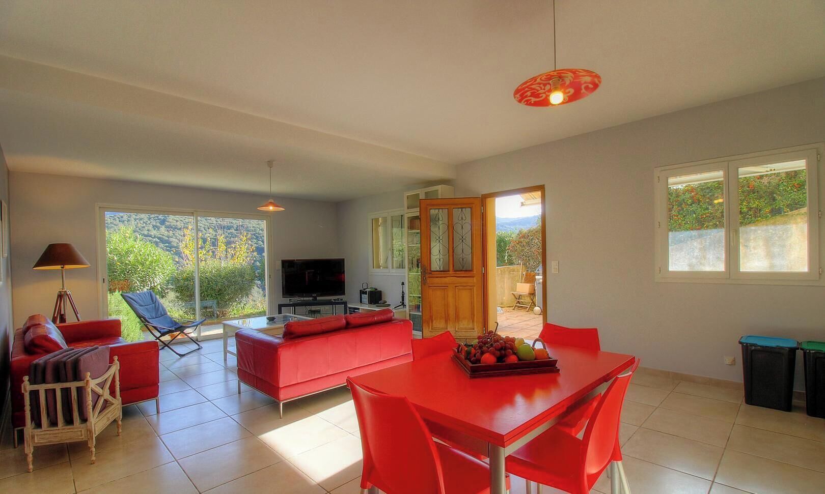 Ein geräumiger Wohnbereich mit einem roten Sofa, einem Esstisch mit roten Stühlen, einem Fernseher und einem großen Fenster mit Blick auf Grünflächen.