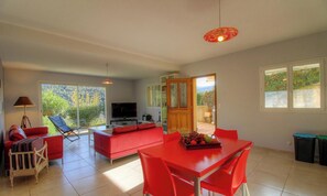 A spacious living area with a red sofa, a dining table with red chairs, a TV, and a large window offering a view of greenery. - Murta - OCANA - Gîte (OCANA)