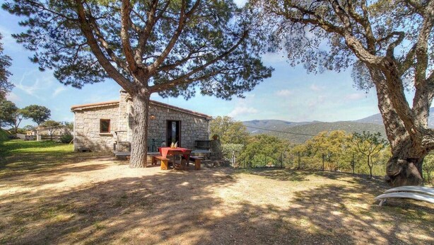 Un bâtiment en pierre avec une table rouge et des bancs, entouré d’arbres et offrant une vue sur des collines lointaines.