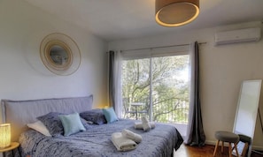 A bedroom with a bed, a round mirror, a window with a view of trees, a small table, and an air conditioning unit. - Casa Chjarina - SOTTA (SOTTA)