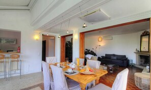 A dining area with a wooden table set for four, surrounded by white chairs. There is a kitchen area with a bar and chairs to the left. A dark-colored sofa and a small round table are in the living area. - Casa Chjarina - SOTTA (SOTTA)