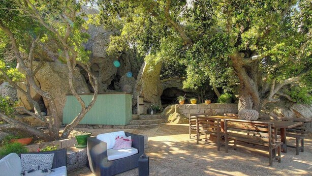 Zona d'estar a l'aire lliure de caràcter rústic, amb taula de fusta i bancs, envoltada d'arbres alts i una paret de roca.