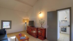 Una sala de estar con un sofá, una mesa de centro, un televisor sobre un mueble de madera, una lámpara y una puerta que da a una cocina.