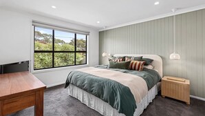 Un dormitorio con un ventanal grande, una cama con muchas almohadas, una mesita de noche y un pequeño aparador de madera.