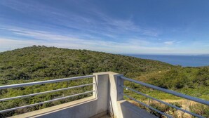 Varanda de concreto com grades de metal, vista para uma paisagem ondulada com vegetação densa e o mar.