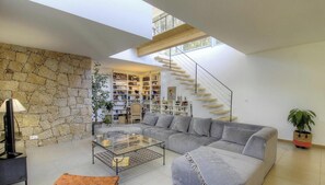 A modern living room with a stone wall, a grey sofa, a glass coffee table, and a staircase leading to an upper level. - Casa Culumbu - SOTTA (SOTTA)