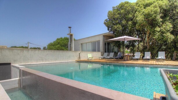 Ein Swimmingpool mit Liegestühlen und einer überdachten Liegefläche, umgeben von Bäumen und einem modernen Gebäude.