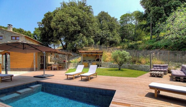 Un patio trasero con piscina, sillas de descanso, zona de barbacoa y un muro de piedra.