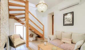 Ein Loft-Zimmer mit freiliegenden Holzbalken, einer Treppe, einer Sofagarnitur, einem Sessel, einer Chandelüster und einem gerahmten Bild an der Wand.