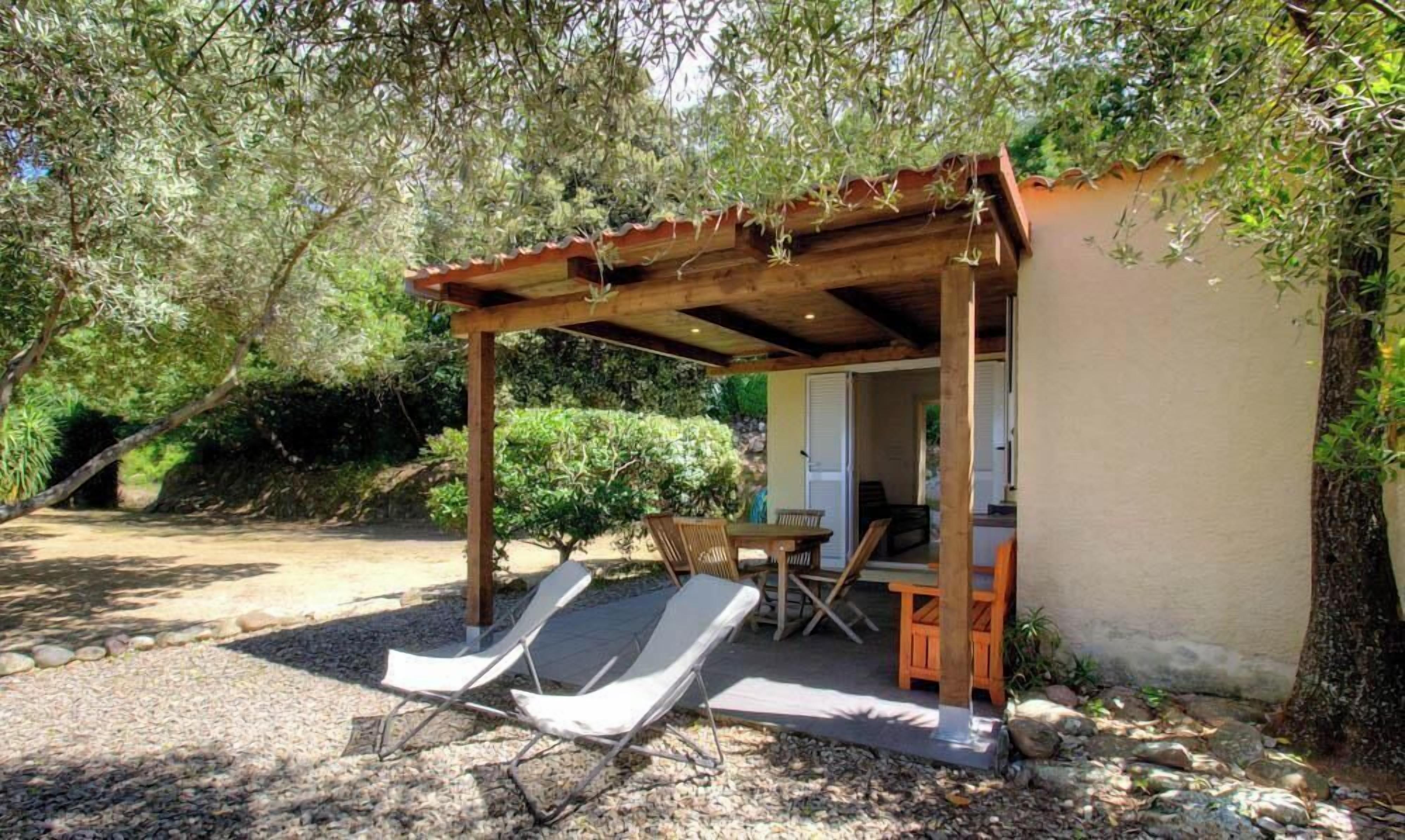 A covered outdoor seating area with a table and chairs, a small structure with a door, and a gravel path.
