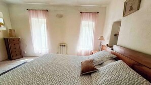 Een slaapkamer met een groot bed, twee ramen met gordijnen, een nachtkastje met lamp en een dressoir.