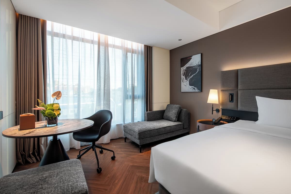 Une chambre d’hôtel avec un lit, un bureau, une chaise, un canapé, une lampe et une fenêtre avec des rideaux.