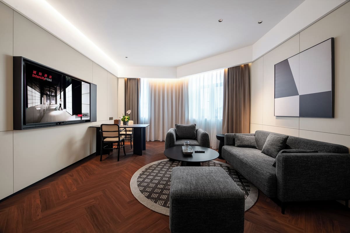 Una habitación de hotel moderna con una televisión de pantalla plana, un sofá, una mesa de centro, un rincón de comedor y ventanas grandes con cortinas.