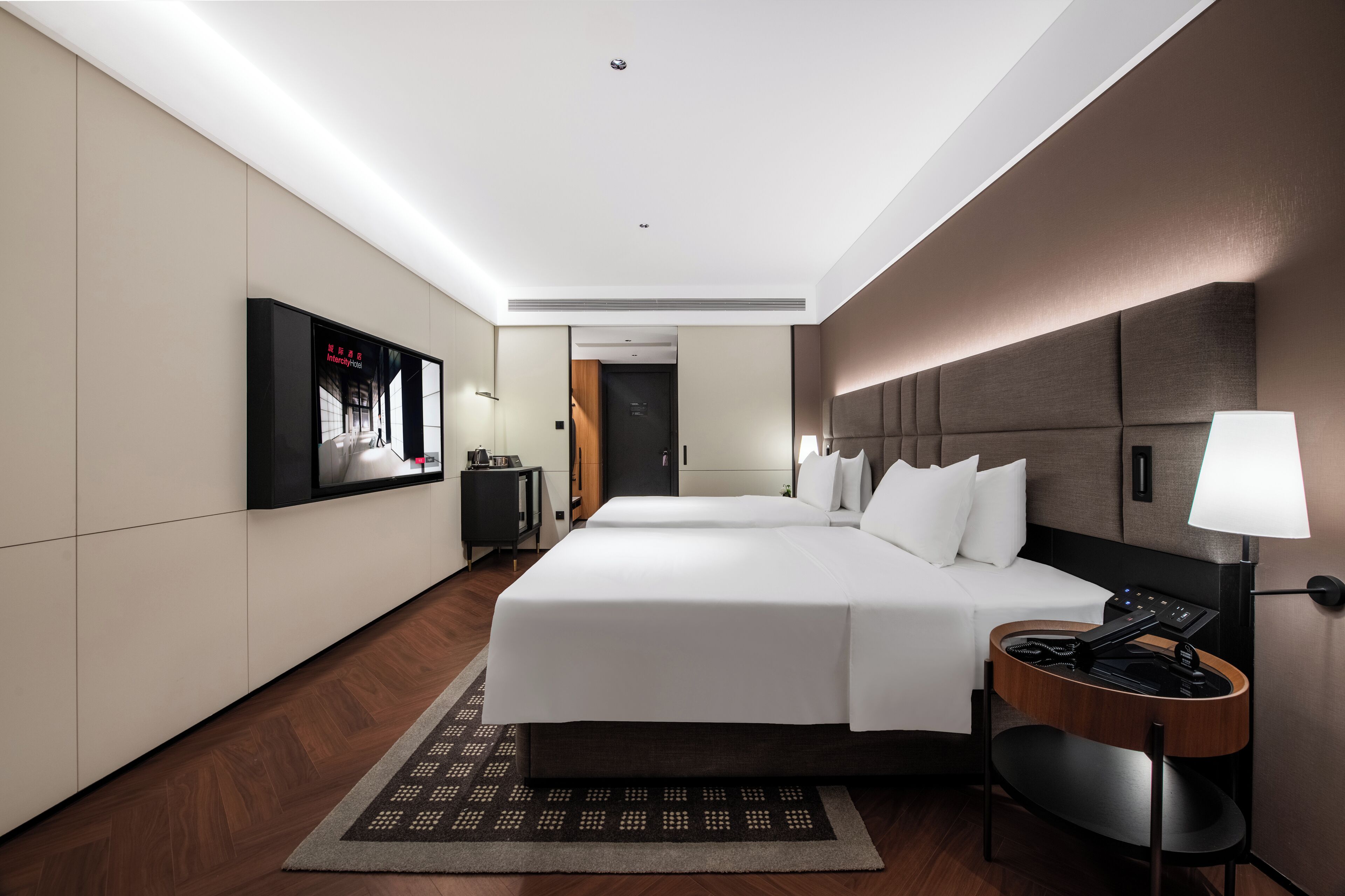 A hotel room with a large bed, a desk with a lamp, a TV mounted on the wall, and a door leading to another room.