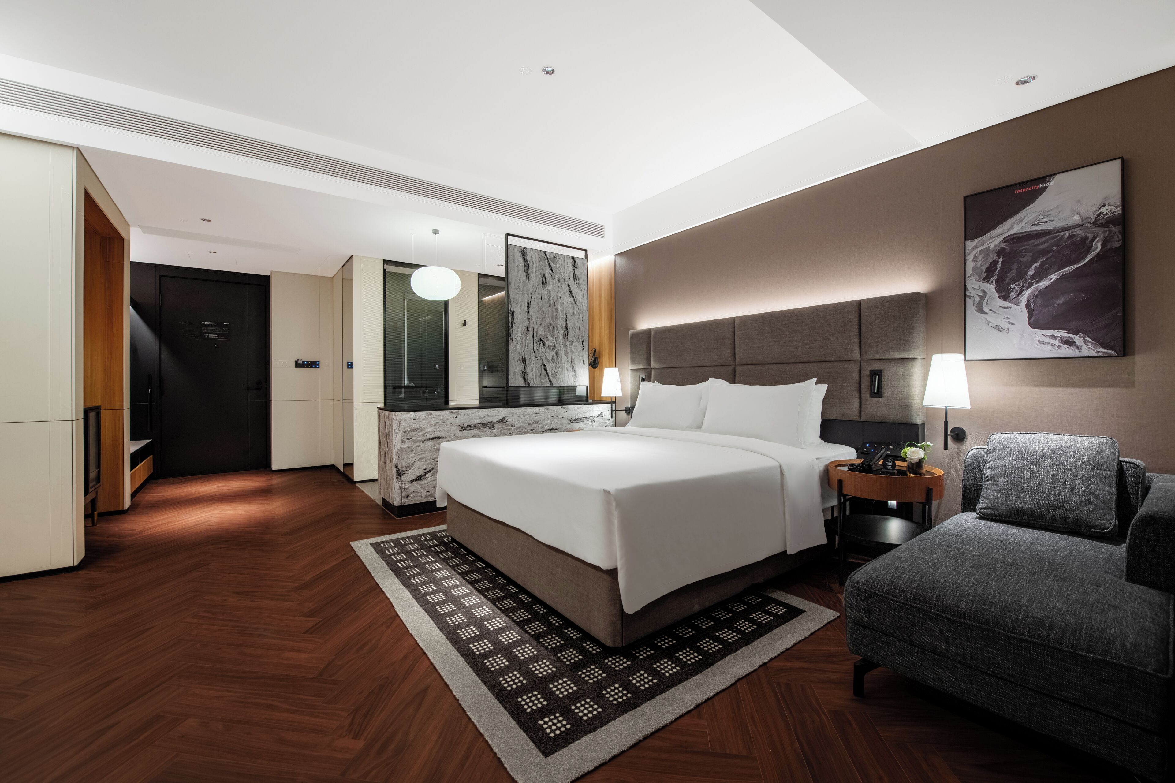 A modern hotel room with a large bed, a desk, a chair, a lamp, and a painting on the wall.