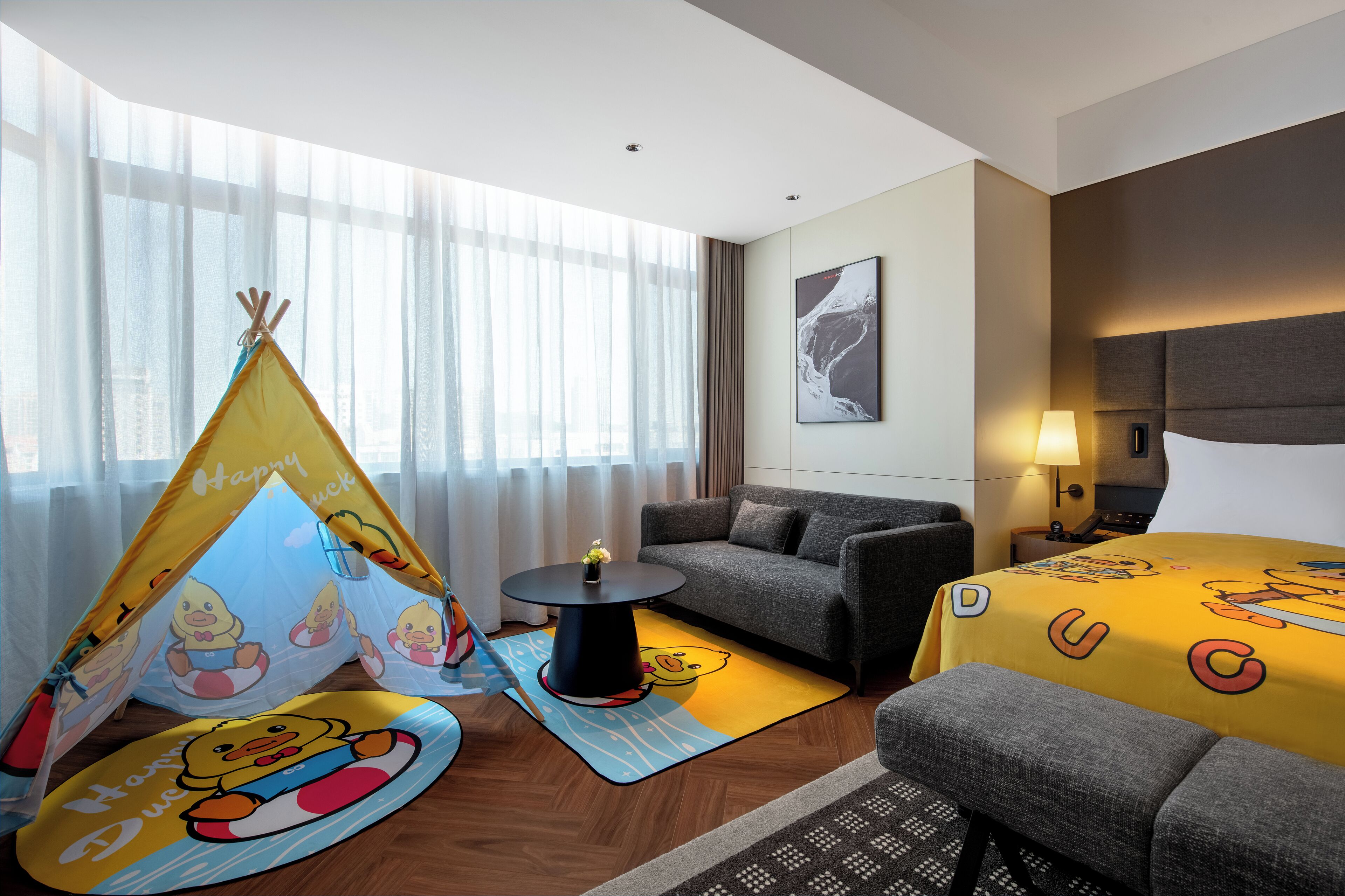 Habitación de hotel con cama, sofá, una mesita con lámpara, ventana con cortinas translúcidas y una tienda de juegos con temática de patos.