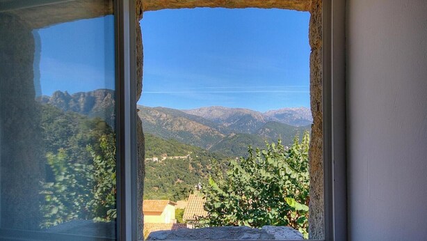 Ein Fenster mit Blick auf eine gebirgige Landschaft, eine steinerne Kante und ein Gebäude mit Ziegeldach.