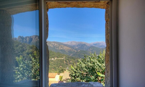 Una ventana con vista a un paisaje montañoso, un alero de piedra y un edificio con techo de tejas.
