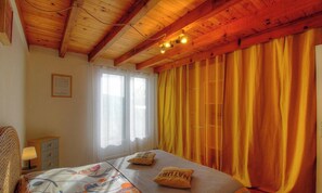 Un dormitorio con techo de madera, una cama con colcha estampada, dos almohadas, una mesita de noche con lámpara y una ventana con cortinas ligeras.